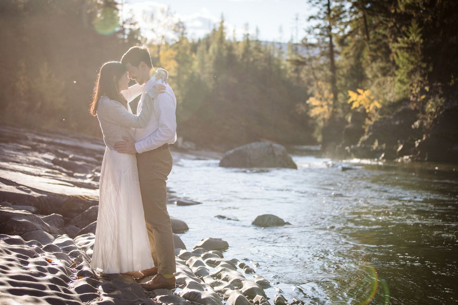Couple standing in sunshine along a riverbank during their Glacier National Park engagement session. 